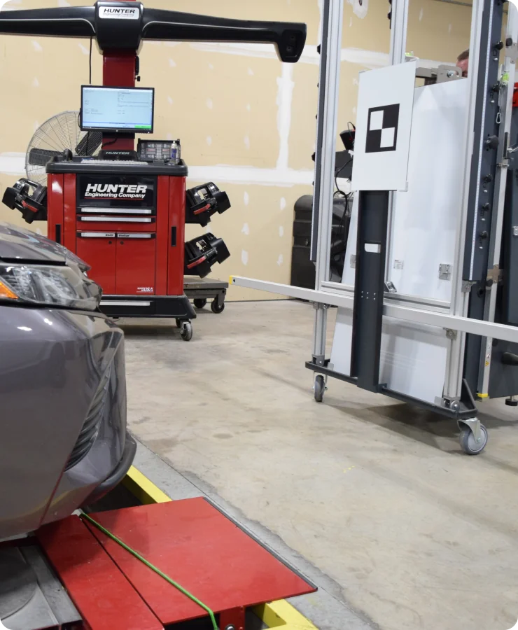 A car is positioned in an auto shop with a Hunter alignment system and calibration equipment set up near its front bumper. A computer screen and tools are visible in the background.