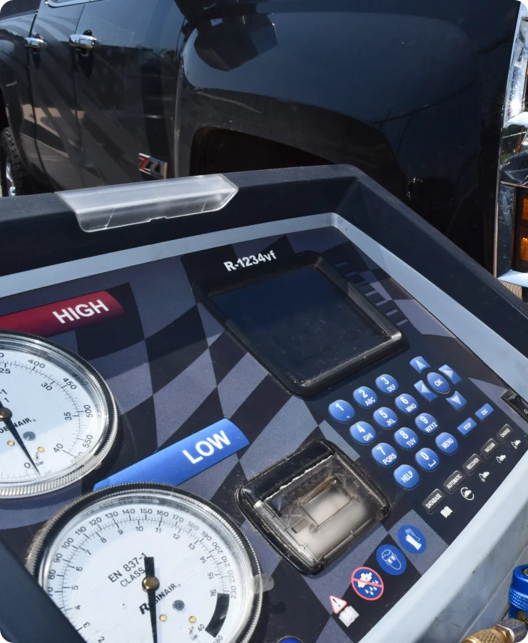 Close-up of an automotive air conditioning service machine showing pressure gauges, buttons, and a display screen, with a black truck visible in the background.
