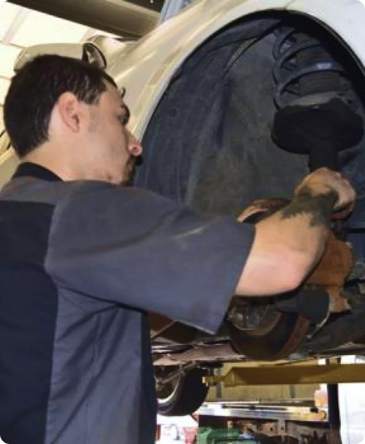 A mechanic wearing a dark uniform works on the suspension and brake system of a car lifted on a service bay. The mechanic is using tools and focusing on the vehicle’s wheel area.