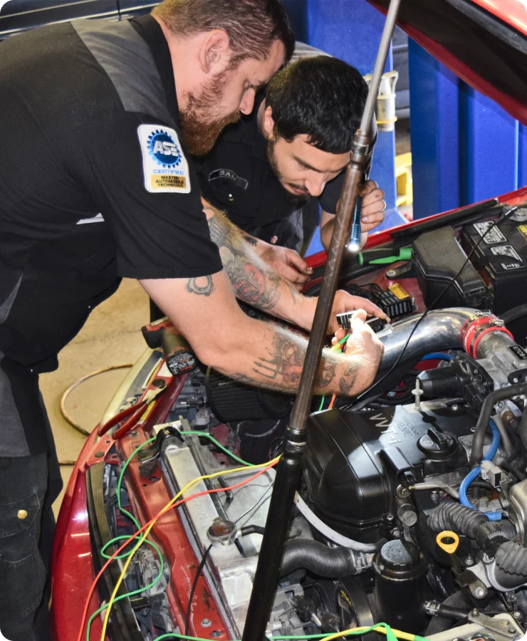 Two mechanics work together under the hood of a red car, examining and repairing wires in the engine bay. One holds tools while the other focuses closely, both wearing black uniforms with an ASE certification patch.