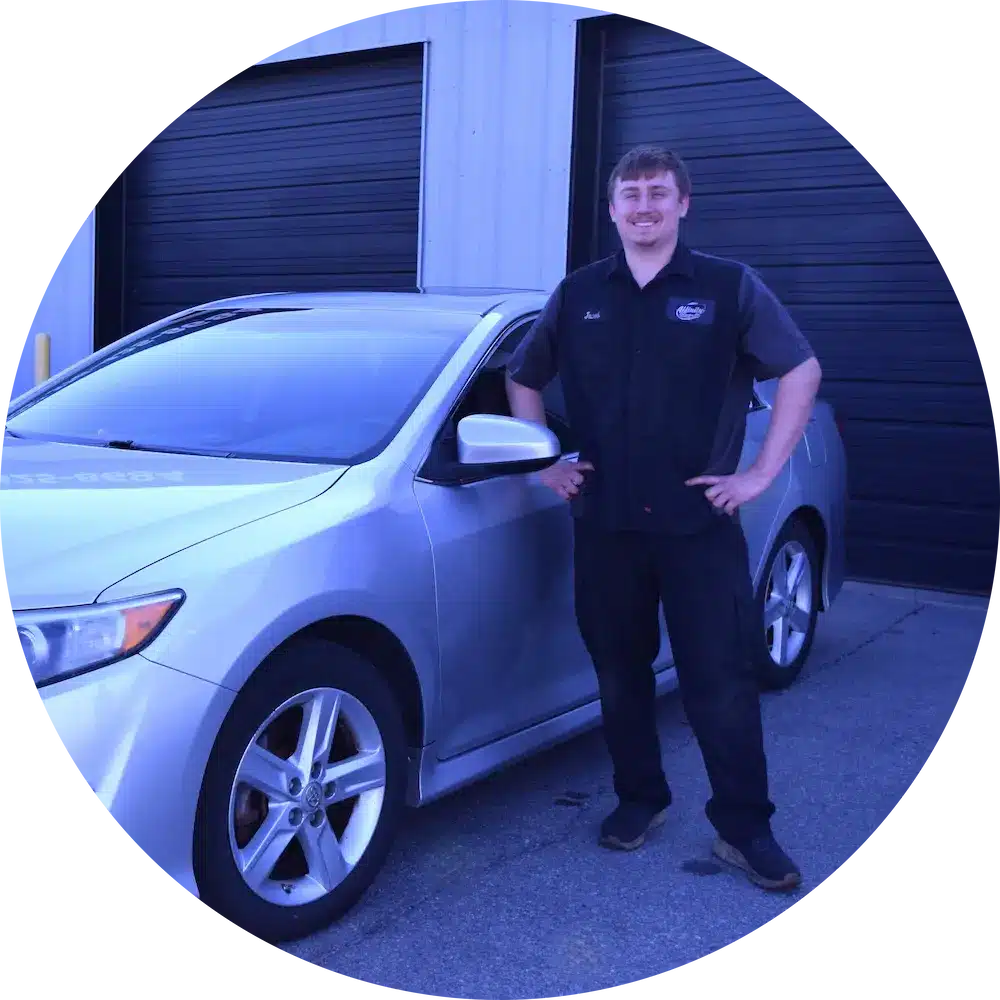 A person wearing a dark uniform stands next to a silver car parked outside a building with black garage doors.