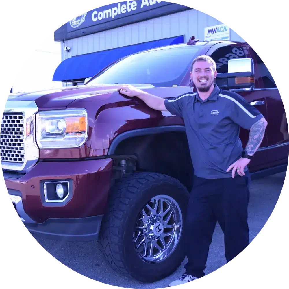 A smiling person in a dark polo shirt stands next to a large maroon pickup truck outside an automotive shop with a blue awning.