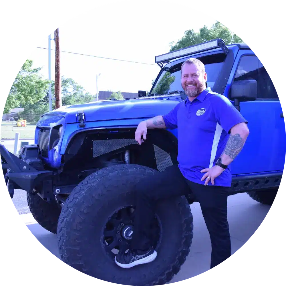 A smiling man with a beard, wearing a blue collared shirt and black pants, leans on the large front tire of a blue off-road vehicle parked outdoors.