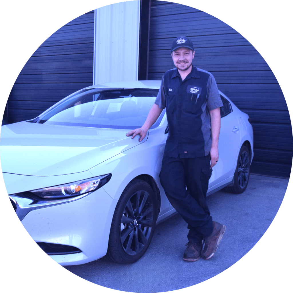 A man in a mechanic uniform and cap stands smiling with one hand resting on the hood of a white car, in front of a black garage door.