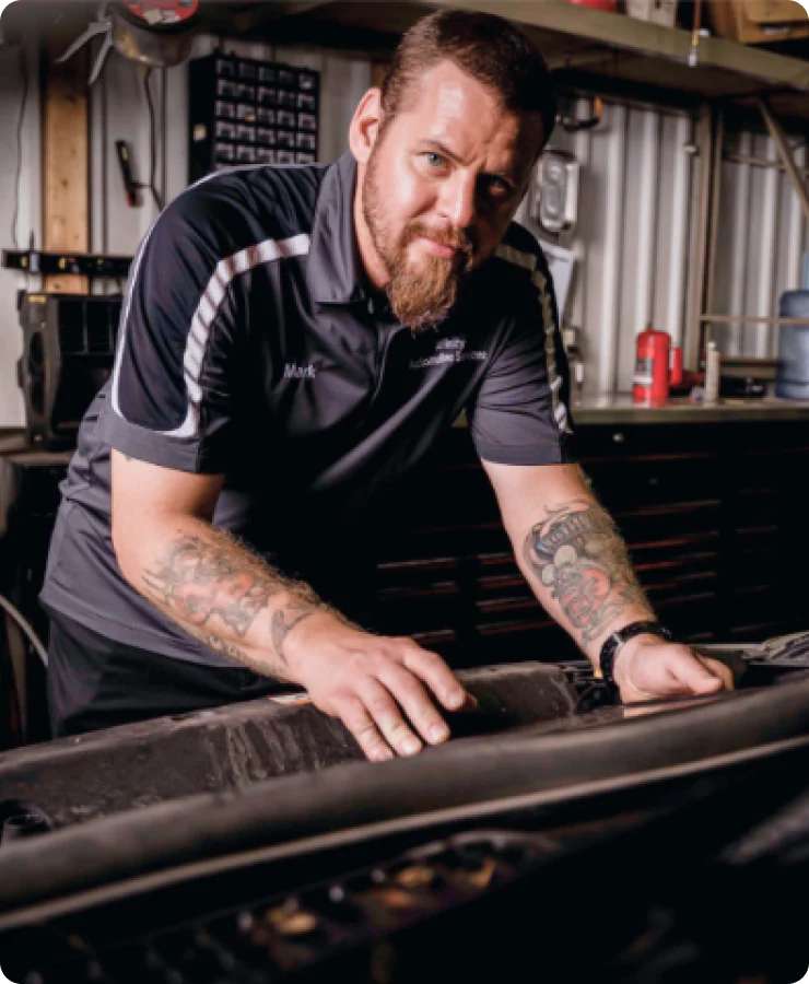 A bearded man with tattoos and a dark polo shirt leans over a car engine in a workshop, looking at the camera. Shelves with automotive tools and supplies are visible in the background.