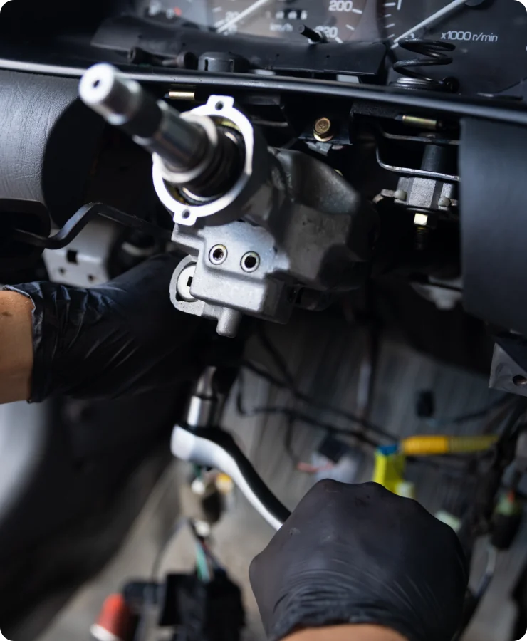 A person wearing black gloves works on a car’s exposed steering column and dashboard, using tools to perform repairs or maintenance.