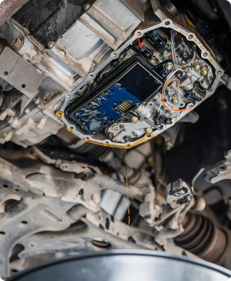 Close-up view of the underside of a vehicle with the automatic transmission oil pan removed, showing exposed transmission components and fluid draining into a container below.