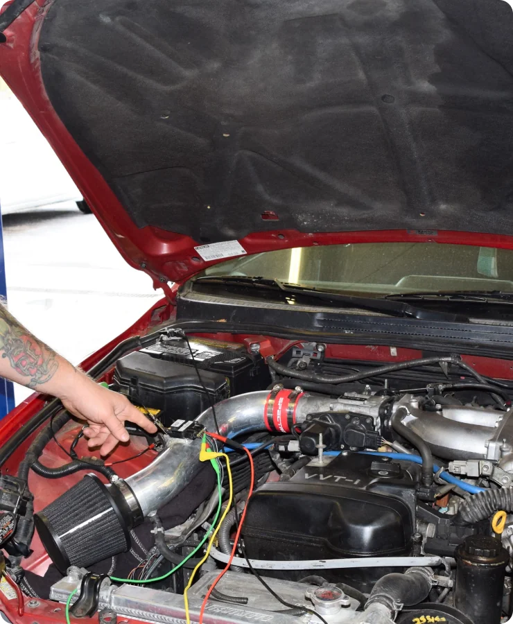 A person with a tattooed arm points to a modified car engine with colored wires and an exposed air intake, under an open hood of a red vehicle.