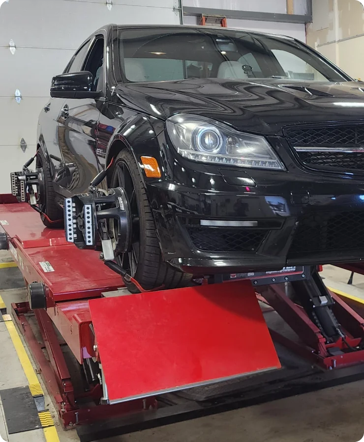 A black car is positioned on a red alignment rack in an auto shop, with alignment sensors attached to its wheels for a wheel alignment procedure. The garage door is visible in the background.