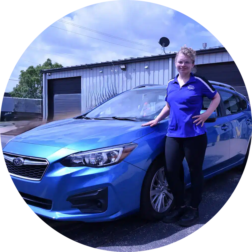 A person wearing a blue polo shirt and black pants stands beside a blue Subaru car, smiling, with one hand resting on the car’s hood. A metal building and a partly cloudy sky are in the background.