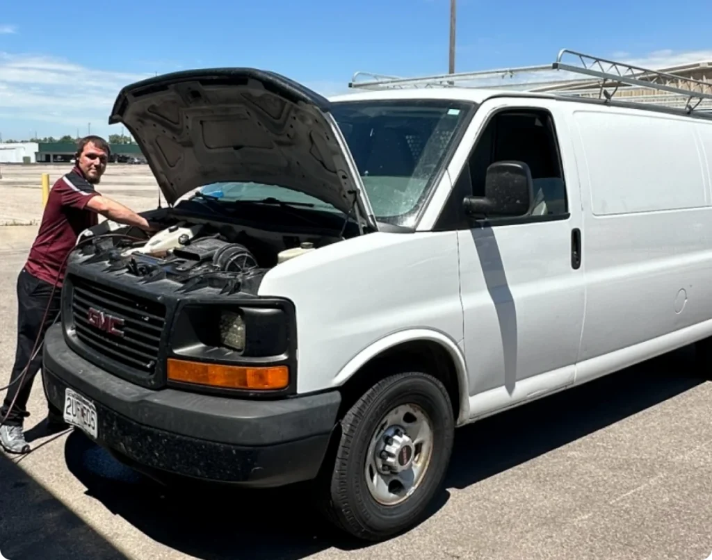 A man wearing sunglasses stands by a white GMC van with its hood open, inspecting the engine in a sunny outdoor parking area. A ladder is secured on the van’s roof.