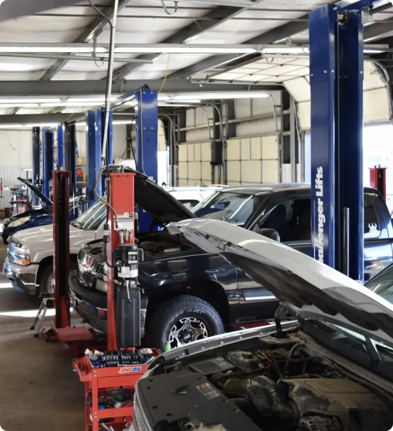 An auto repair shop with several vehicles parked inside, many with their hoods open, and blue vehicle lifts along the work bays. Various tools and equipment are visible throughout the garage.