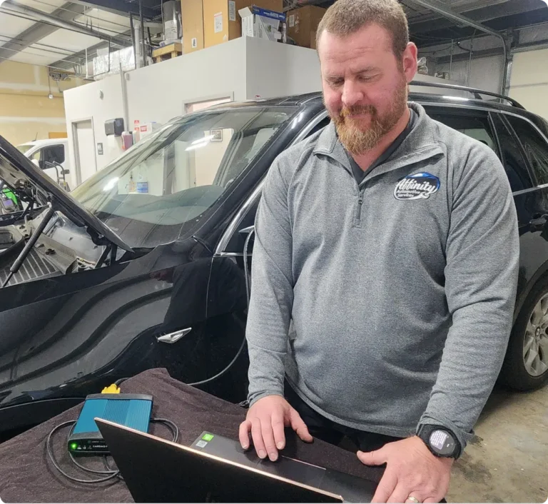 A man in a gray pullover stands next to a black car with its hood open, working on a laptop. Auto repair equipment is visible on the table in a well-lit garage or workshop.