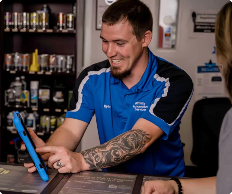 A man in a blue Affinity Automotive Service shirt smiles while showing a tablet to a customer across the counter. Shelves with automotive products are visible in the background.