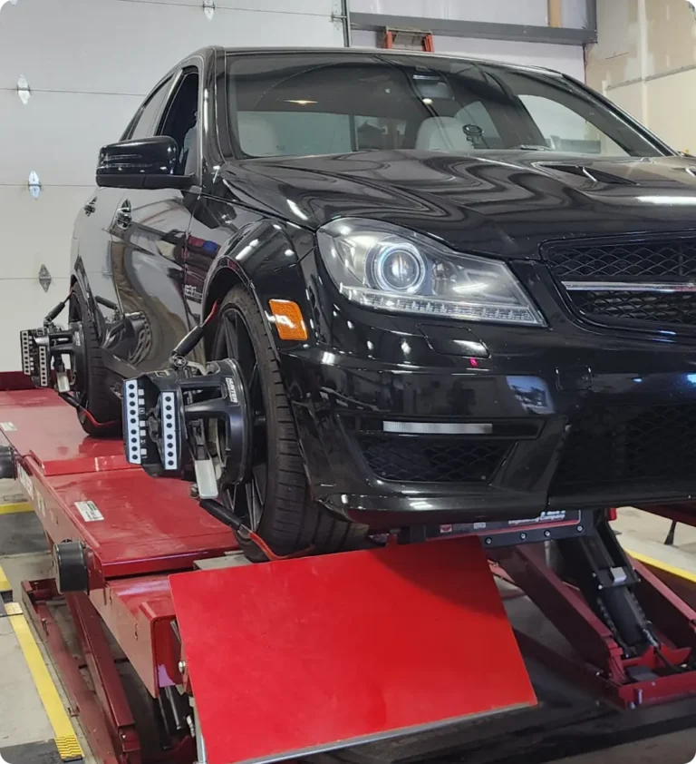 A black sports car is on a red platform in an auto repair shop, with wheel alignment equipment attached to its front wheels. The shop has a white garage door in the background.