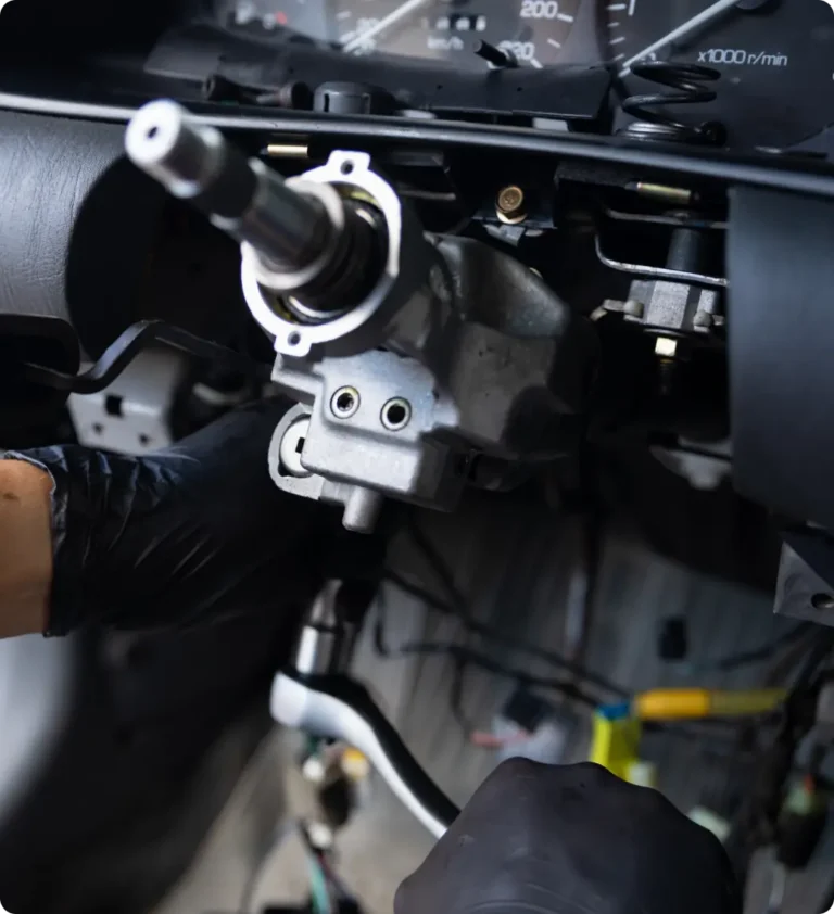 A person wearing black gloves works on the exposed steering column of a car, with dashboard panels removed and wires visible.