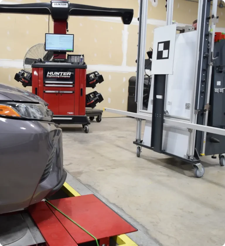 A car in an auto repair shop undergoing alignment calibration with a Hunter alignment machine and a target board for ADAS calibration in the background.