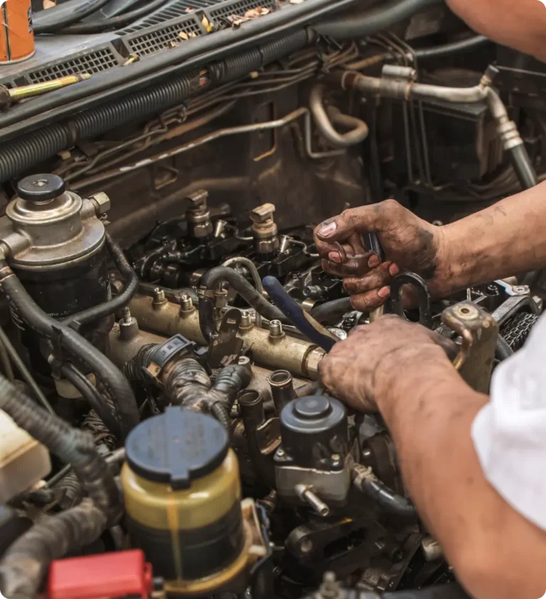 A mechanic with dirty hands uses a wrench to work on a car engine, surrounded by various engine components and tools under the vehicle’s hood.