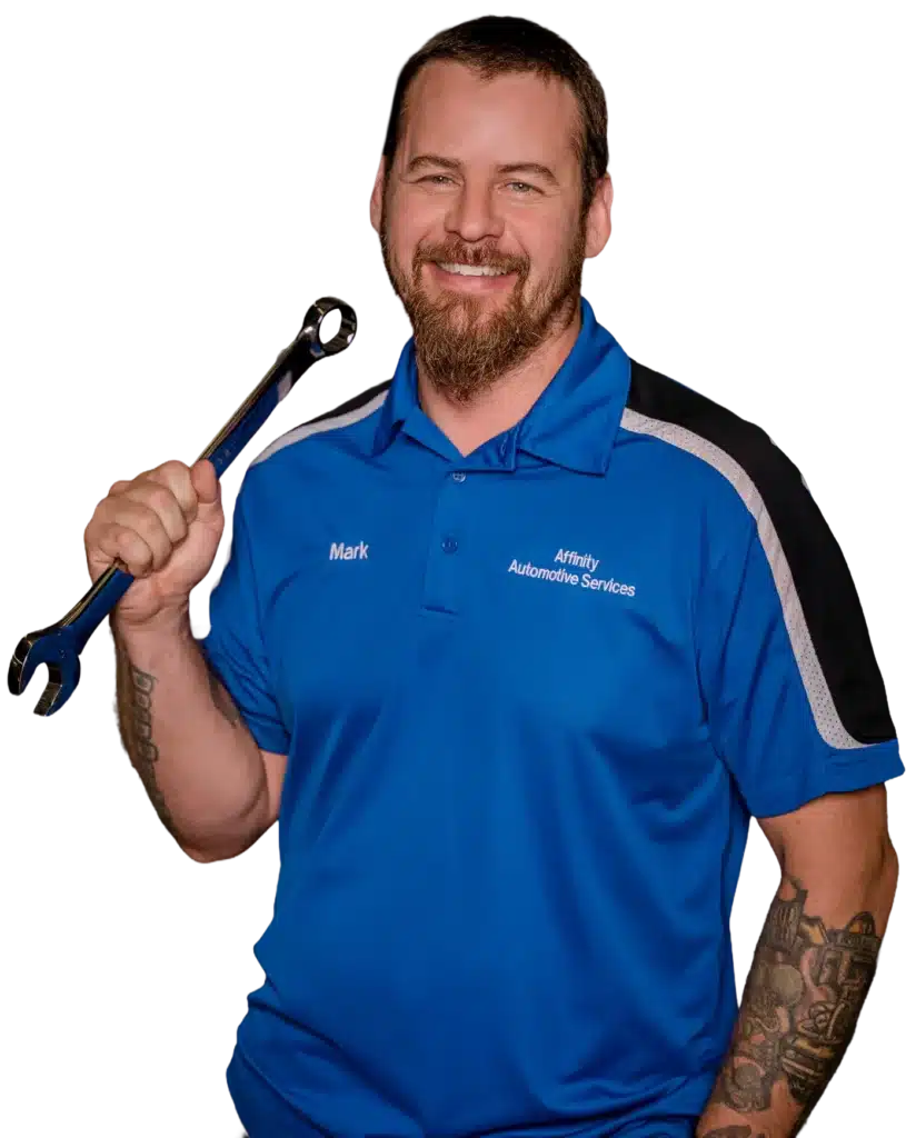 A smiling man with a beard wearing a blue Affinity Automotive Services polo shirt holds a large wrench over his shoulder. He has tattoos on his arms and looks friendly and approachable.