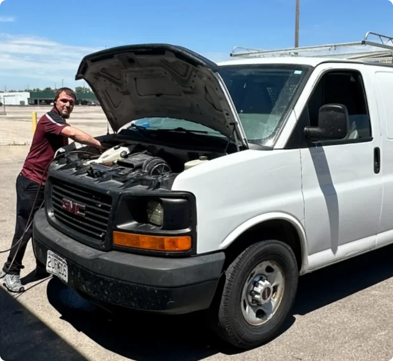 A person stands beside a white GMC van with the hood open in a sunny parking lot, appearing to work on the engine. The individual is wearing a maroon shirt and black pants.