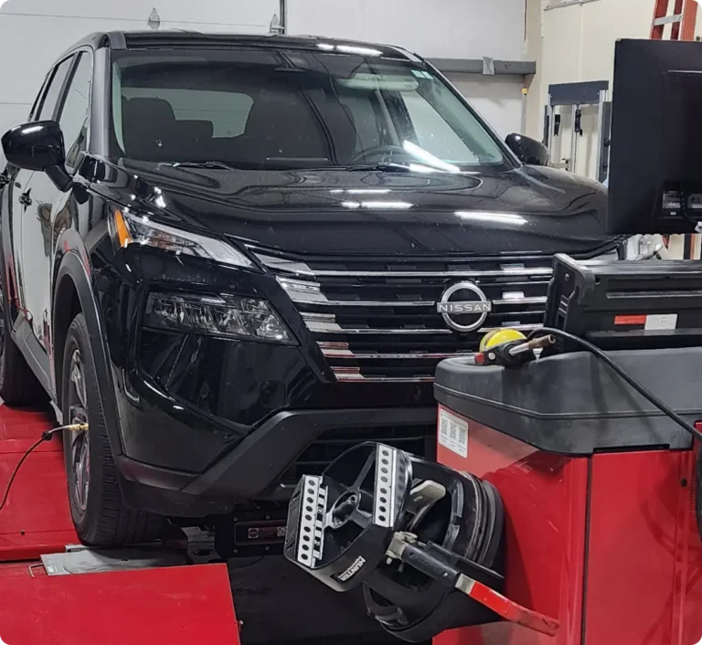 A black Nissan SUV is parked inside an auto repair shop, positioned on a red platform with wheel alignment equipment attached to the front left tire. A computer and diagnostic tools are set up nearby.