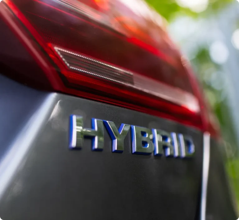 Close-up of a car’s rear with a red taillight and the word “HYBRID” in chrome letters, indicating it is a hybrid vehicle; background is blurred with some greenery visible.