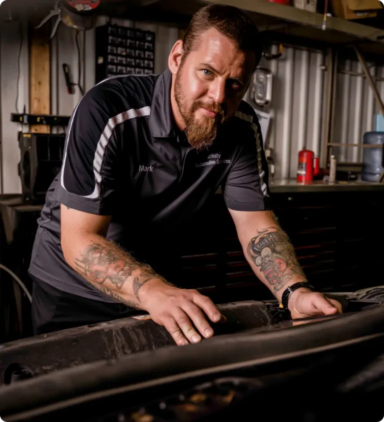 A bearded man with tattoos, wearing a black and gray polo shirt, leans over a car engine in an automotive workshop, looking at the camera. Shelves and tools are visible in the background.