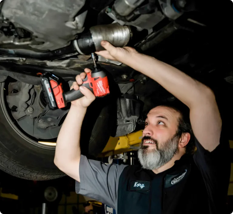 A mechanic with a beard uses a red power drill to repair the underside of a vehicle in an auto shop, looking up while working.