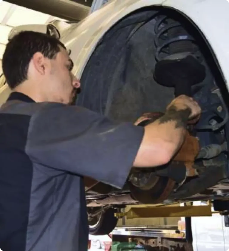 A mechanic works on the suspension and brakes of a raised vehicle in an auto repair shop, using tools to make adjustments inside the wheel well.