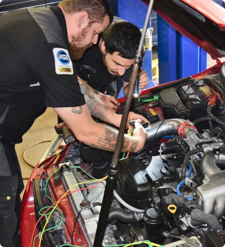 Two auto mechanics work together under the hood of a red car, using tools and examining engine components with colorful wires visible; both wear black uniforms and have tattoos on their arms.