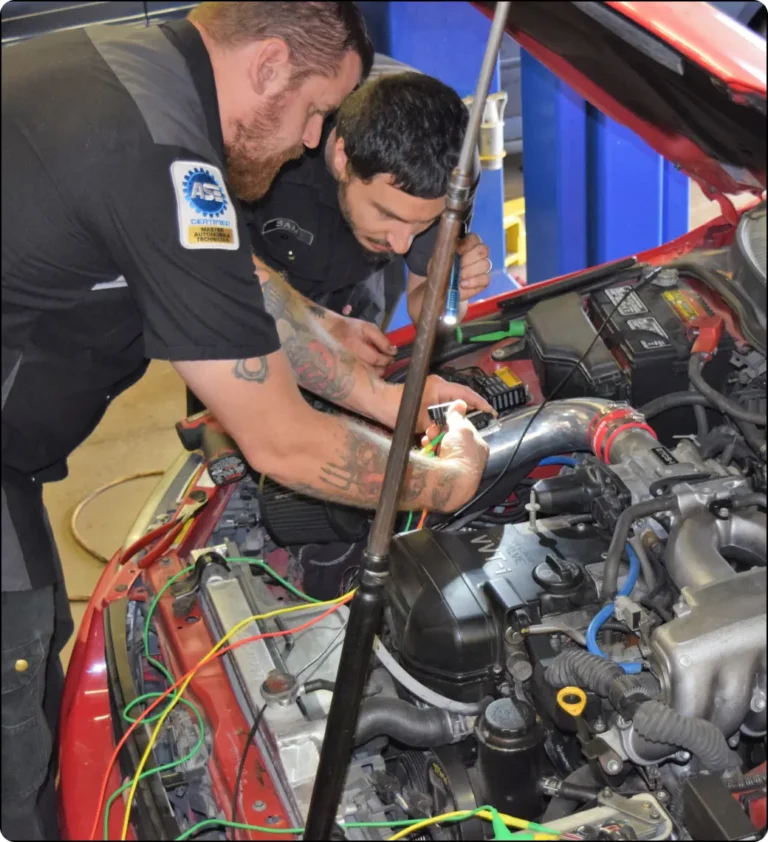 Two auto mechanics work together under the hood of a red car, examining wires and engine components in a garage setting. One has tattoos and an ASE patch on his sleeve.