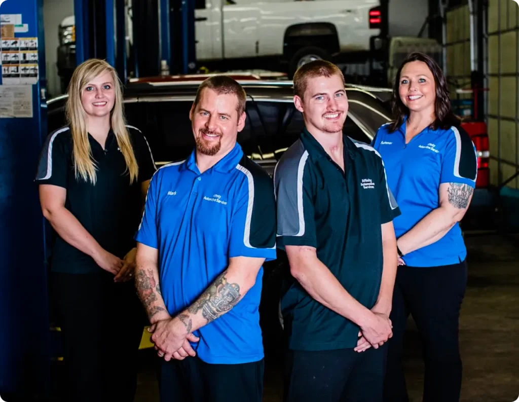 Four smiling auto shop employees, two men and two women, stand in front of a vehicle in a garage. They wear shop uniforms and pose with hands clasped, surrounded by automotive equipment in the background.