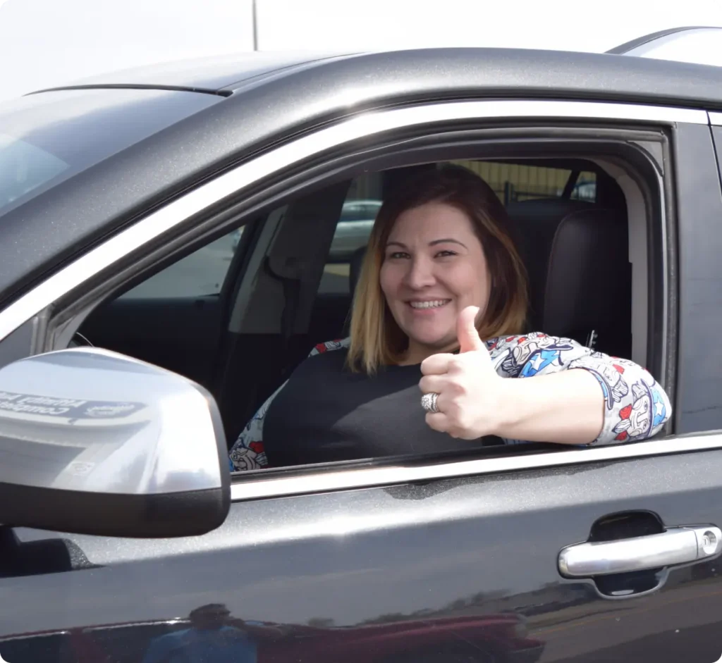 A smiling woman sits in the drivers seat of a car with the window down and gives a thumbs-up gesture.