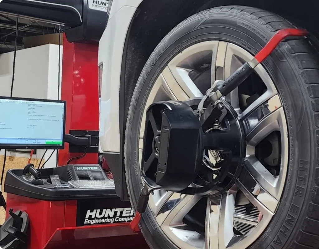 A close-up of a car wheel on an alignment machine, with sensors attached to the rim and a computer displaying vehicle alignment data in a workshop setting.