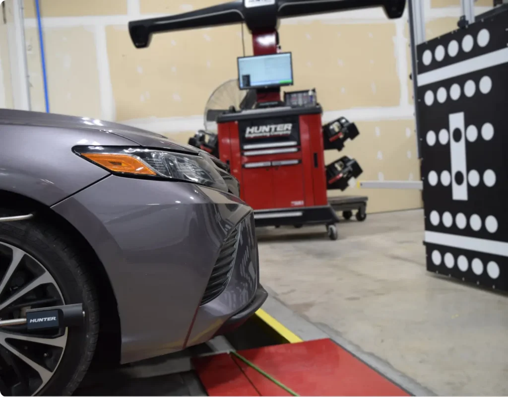 A gray car is positioned in front of a wheel alignment machine in an automotive workshop, with a Hunter alignment system and calibration targets in the background.