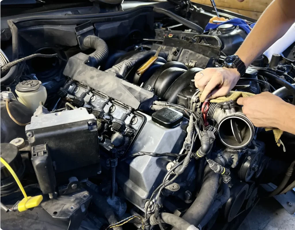 A person works on a car engine, using tools to adjust parts near the throttle body, with wires and engine components visible.