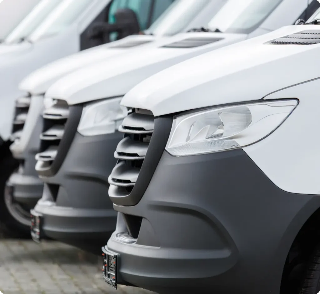 Close-up view of the front ends of several white delivery vans parked in a row, showing their headlights, grilles, and bumpers.