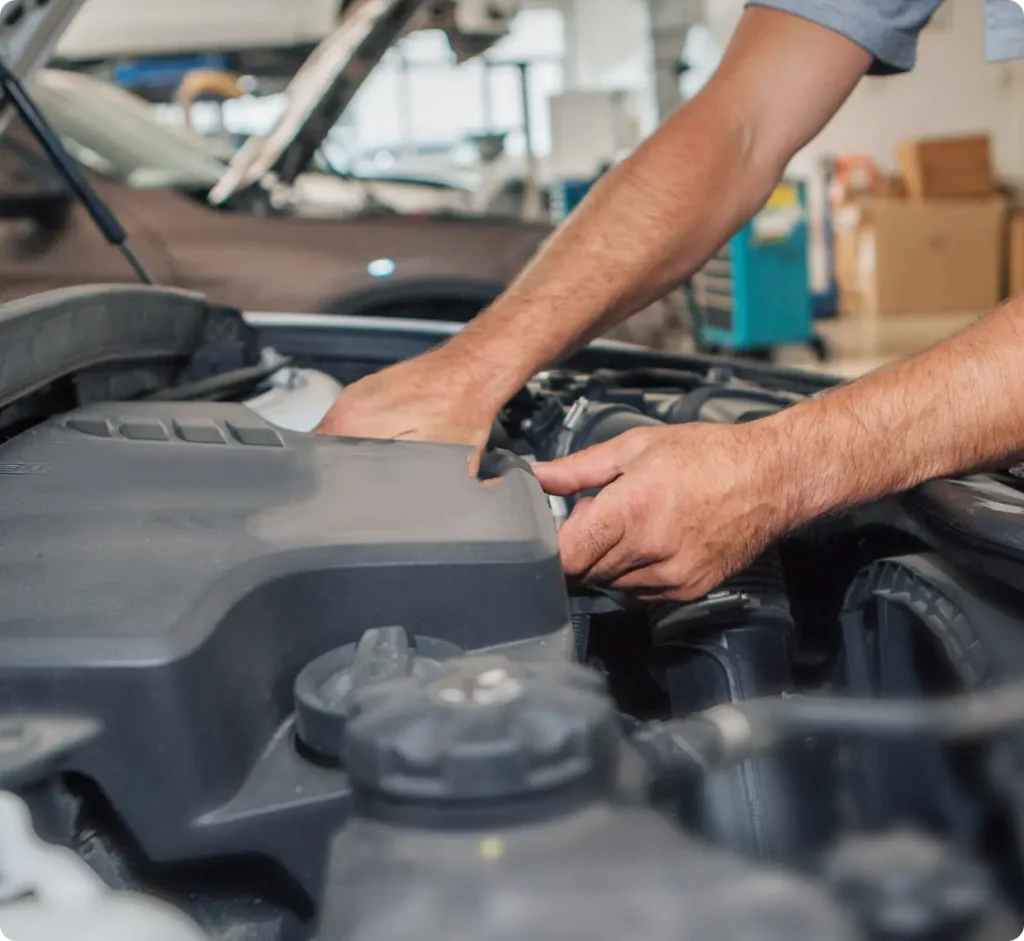 A person’s hands are working under the hood of a car, performing maintenance or repairs in a well-lit garage or workshop.