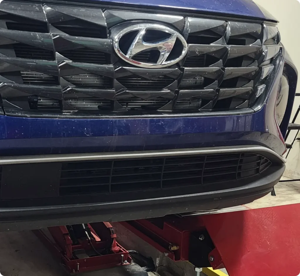 Close-up of the front grille and bumper of a blue Hyundai vehicle, elevated on a red hydraulic lift inside an automotive workshop.