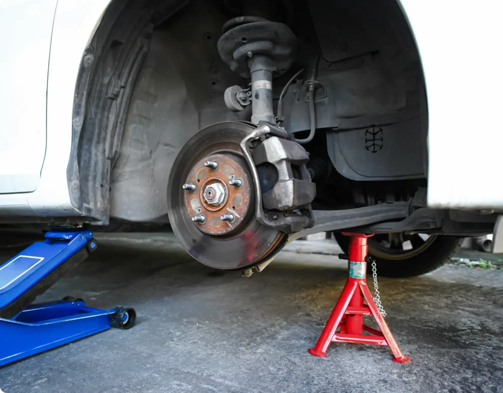 A car with its front wheel removed is supported by a red jack stand and a blue hydraulic floor jack, exposing the brake disc and suspension components.