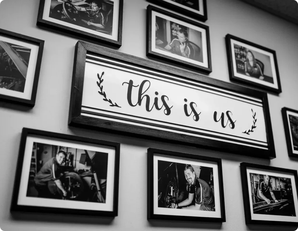 A wall display featuring a large framed sign that reads “this is us,” surrounded by black-and-white photos of people working on cars and smiling, all in rectangular frames.