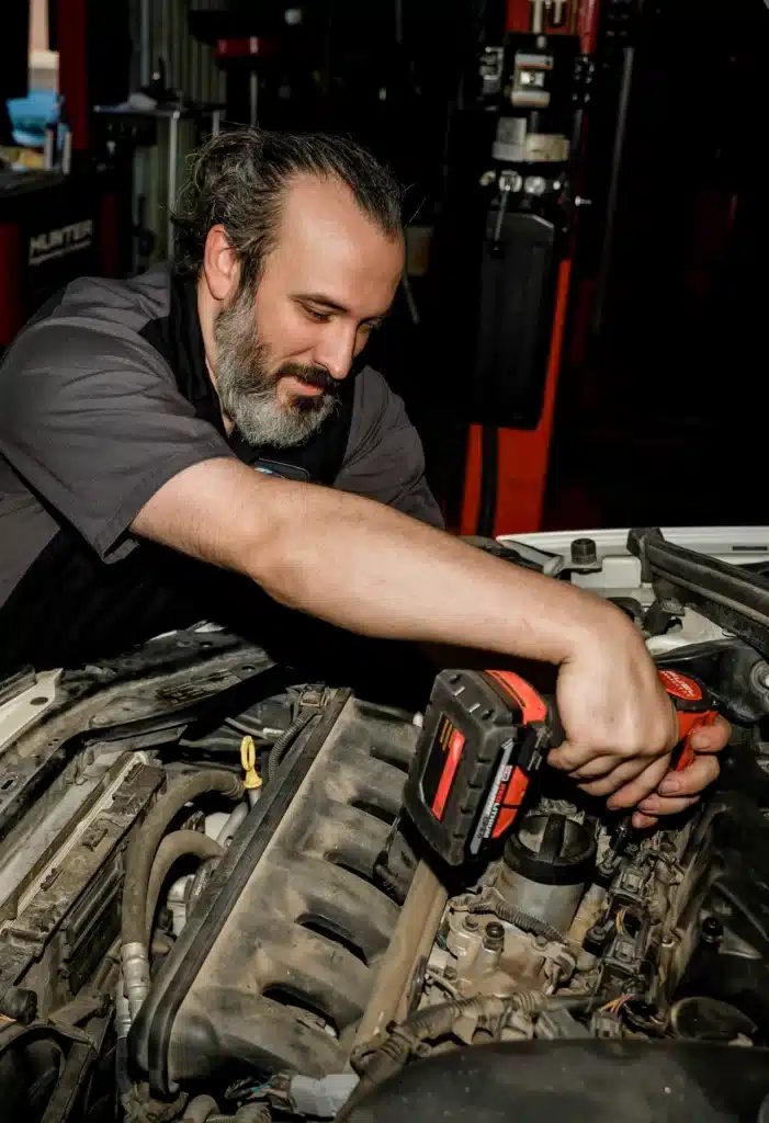 A mechanic with dark hair and a beard uses a power tool to work on a car engine in a garage, focusing intently on the task.