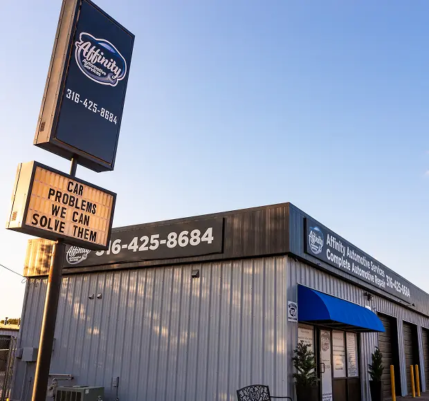 A metal auto repair shop with Affinity Automotive Services signs and a marquee reading CAR PROBLEMS WE CAN SOLVE THEM outside, under a clear sky. The building features large windows and a blue awning above the door.