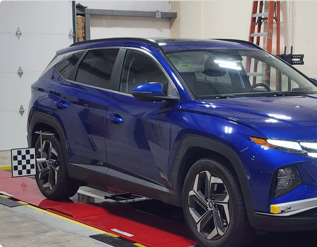 A blue SUV is parked indoors on a red platform, next to a black-and-white calibration board. A ladder is visible against the wall in the background.