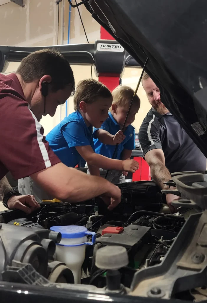Two adults and two young boys look under the hood of a car in a garage. The adults are guiding the boys, who are reaching toward the engine, appearing curious and engaged.