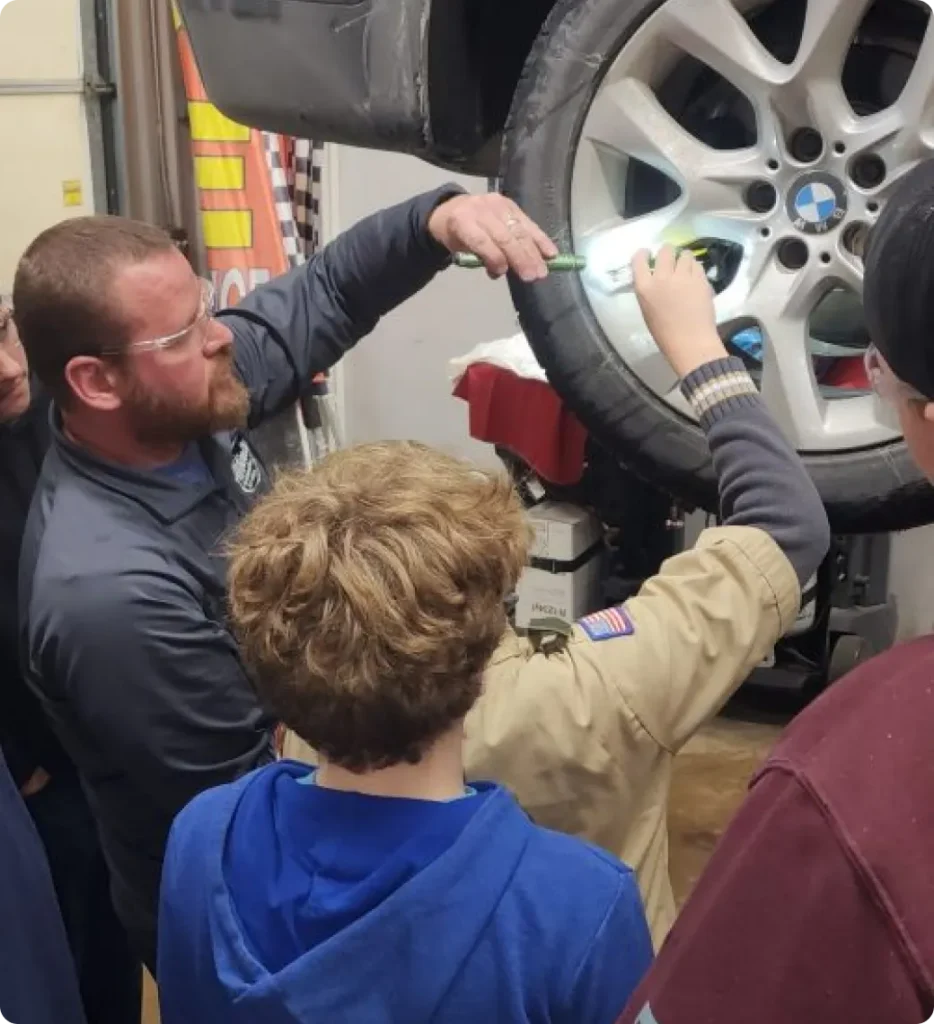 A mechanic explains a car’s tire and wheel to a group of kids, including one in a scout uniform, in an auto shop. The group observes closely, with the mechanic using a tool to point at the tire.