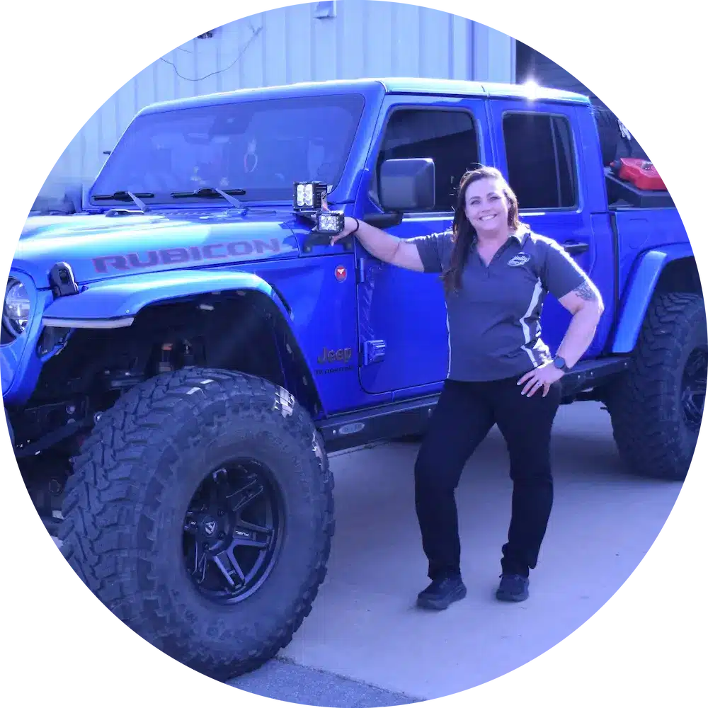 A smiling woman stands next to a lifted blue Jeep Rubicon, resting one arm on the hood, in front of a building. She wears a gray polo shirt, black pants, and black shoes.