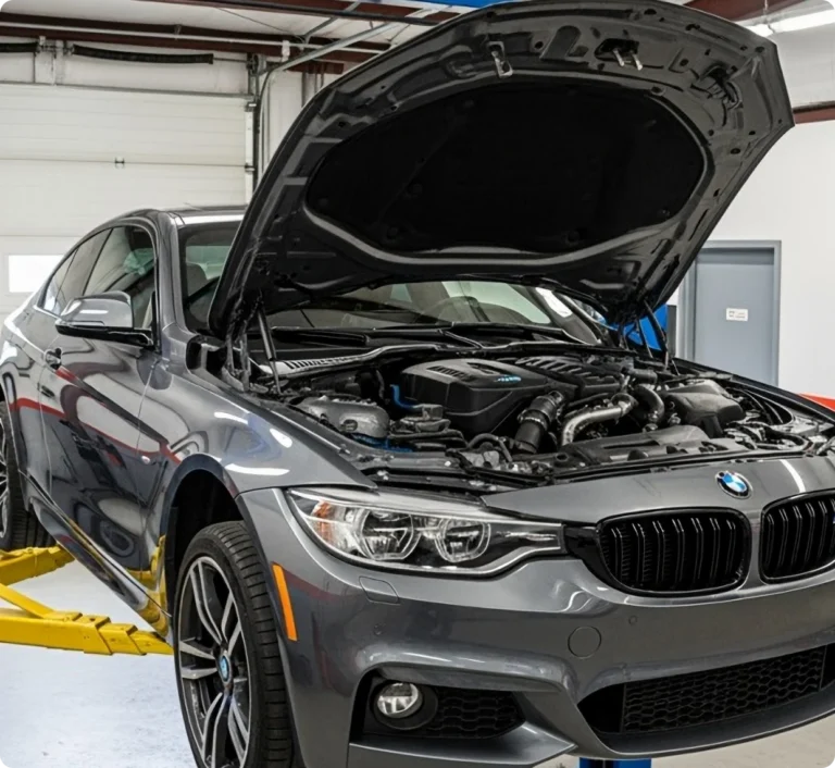 A gray BMW car with its hood open is parked inside a garage on a hydraulic lift, exposing the engine for maintenance or repair.