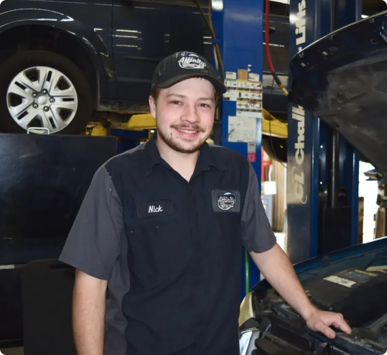 A smiling auto mechanic in a uniform and cap stands beside a car with its hood open in a garage. Another car is lifted on a hydraulic lift in the background.