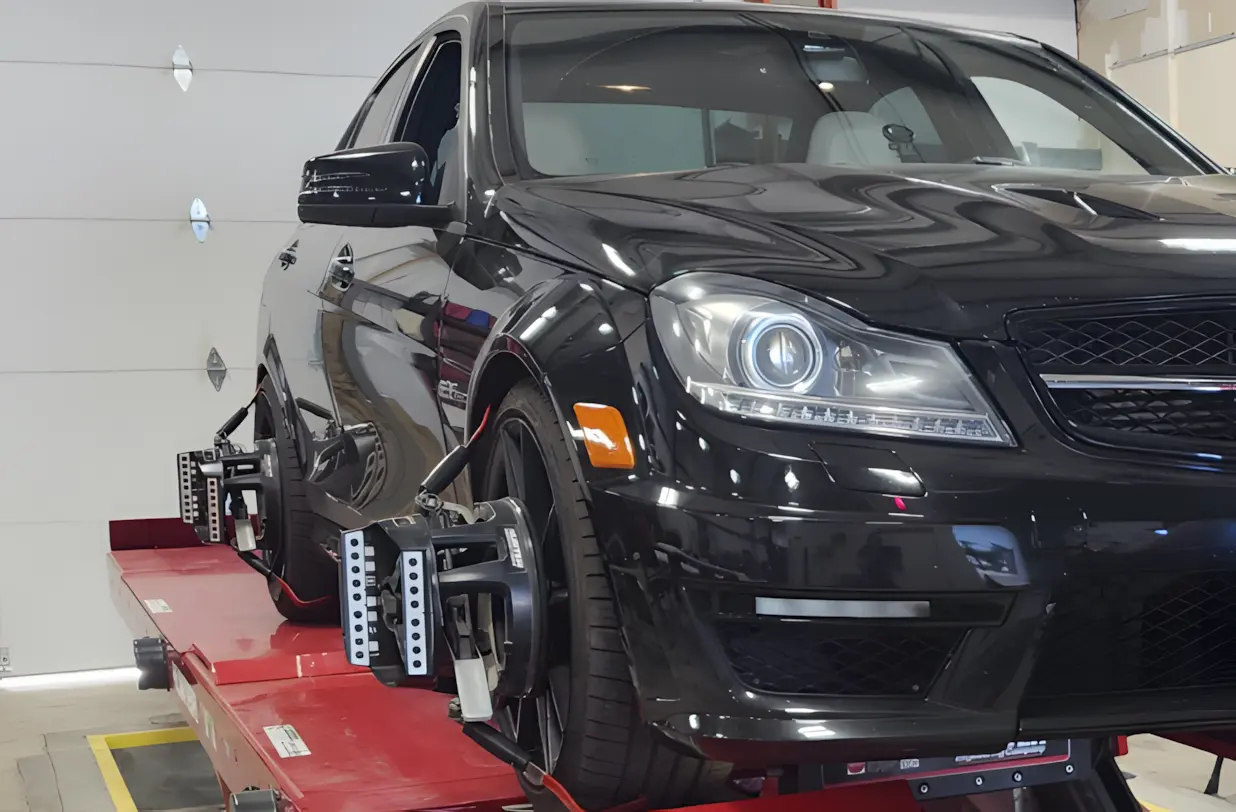 A black car is positioned on an alignment rack in a garage, with sensors attached to each wheel for wheel alignment service.
